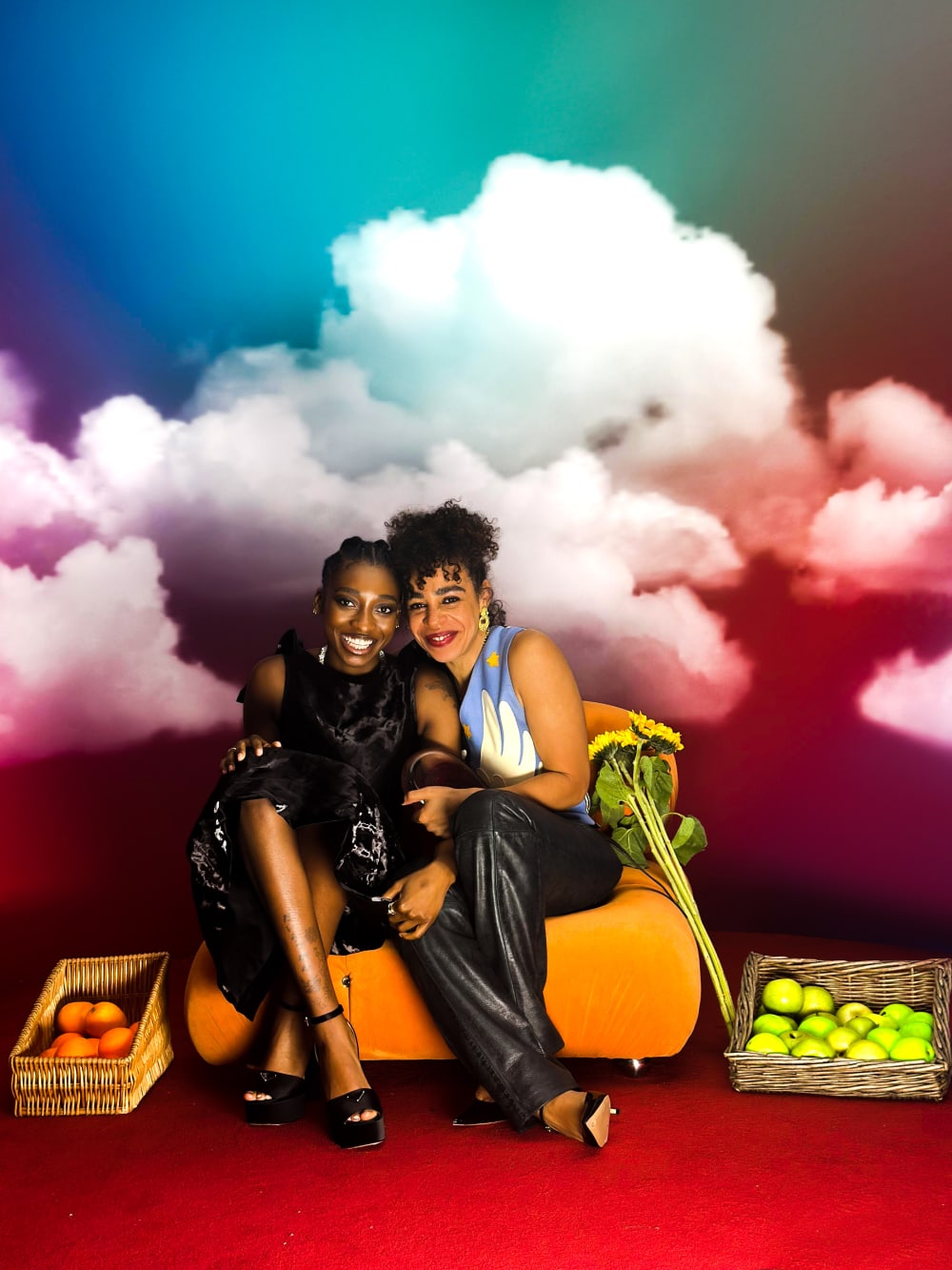 Little Simz and Martine sat on a small sofa - their arms around each other, smiling. The background is high-contrast and there's baskets of fruit on the floor