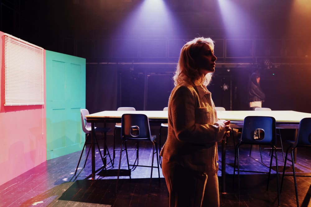 Image shows a person dressed in a white suit standing in a theatre with multicoloured doors and a long table with chairs in the background