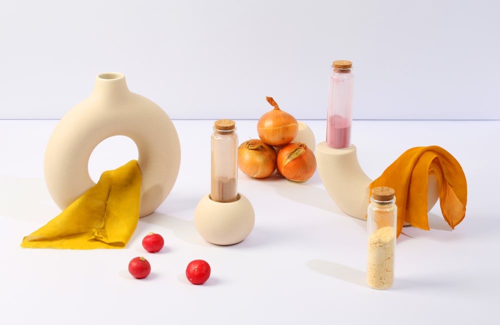 3 corked tubes of dye powders surrounded by beige ceramics, coloured pieces of fabric, onions and fruit. 