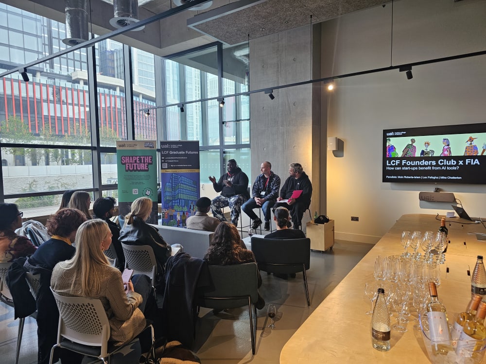 a collection of students in a classroom space, with floor to ceiling windows, listening to 3 speakers at the front of the room on high stools. 