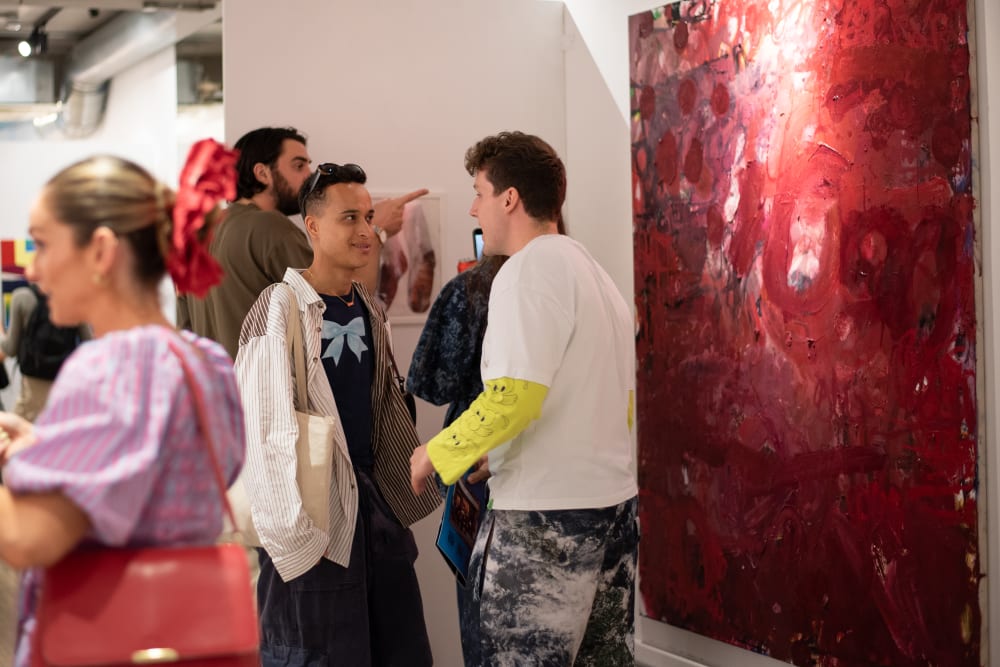 Visitors to the gallery standing and chatting. To the right of the photo, a large red oil painting is on display. 