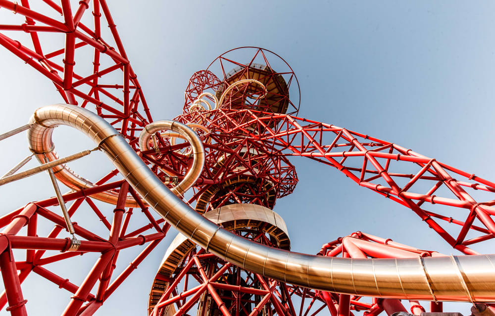 Upward view of the ArcelorMittal Orbit sculpture and slide in Queen Elizabeth Olympic Park, Stratford, London. The red lattice steel structure twists dramatically against a clear blue sky, with the world’s longest tunnel slide spiraling around the tower in shiny metallic tubes. The perspective emphasizes the futuristic design, bold architectural engineering, and dynamic curves of this iconic East London landmark. Keywords: ArcelorMittal Orbit slide, Queen Elizabeth Olympic Park, Stratford London attraction, Olympic sculpture tower, red steel lattice structure, London adventure activities, tallest sculpture UK.