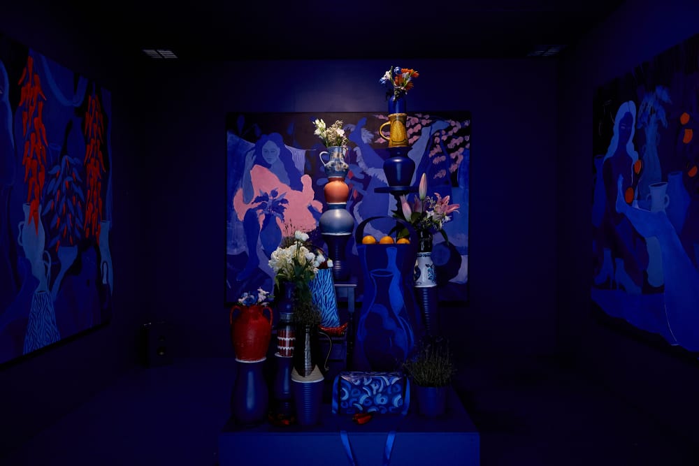 A dark blue room with a painting on each wall. The painting on the back wall is blue and pink and a vase of flowers and a woman are visible. In the foreground is a collection of ceramics, fruit and fresh flowers arranged together on painted blue plinths.
