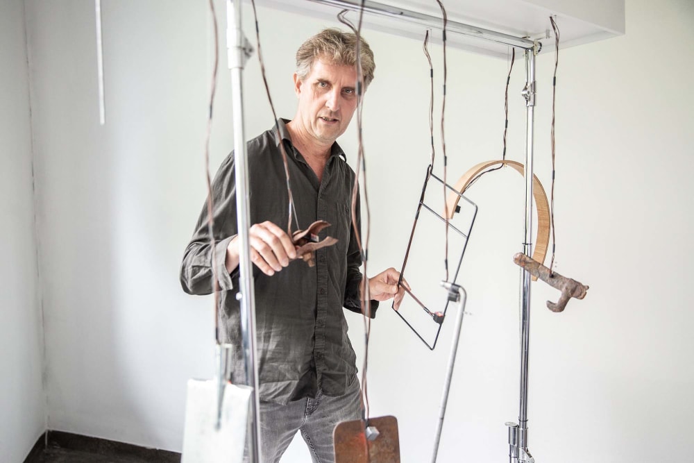 A man in a grey shirt stands behind various objects suspended from the ceiling with electrical wire.