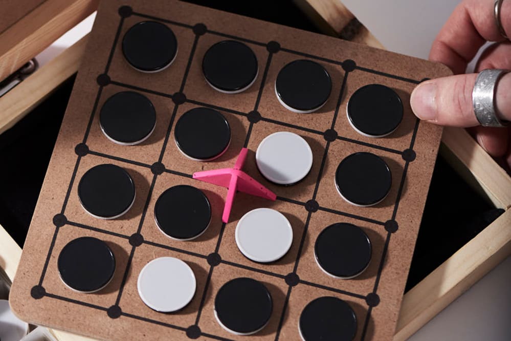 A hand holding a small boardgame with black and white tiles and a pink pointer over a wooden display box