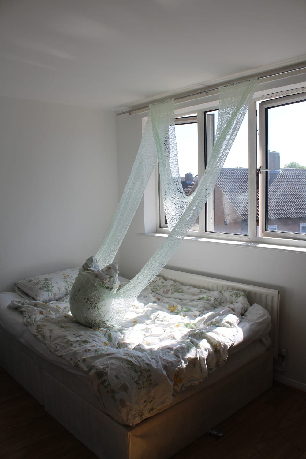A bedroom, a bed pushed up against the window, with sunlight shining through the window onto the bed. A sculpture piece attached to the top of the windowns hanging down onto the bed.