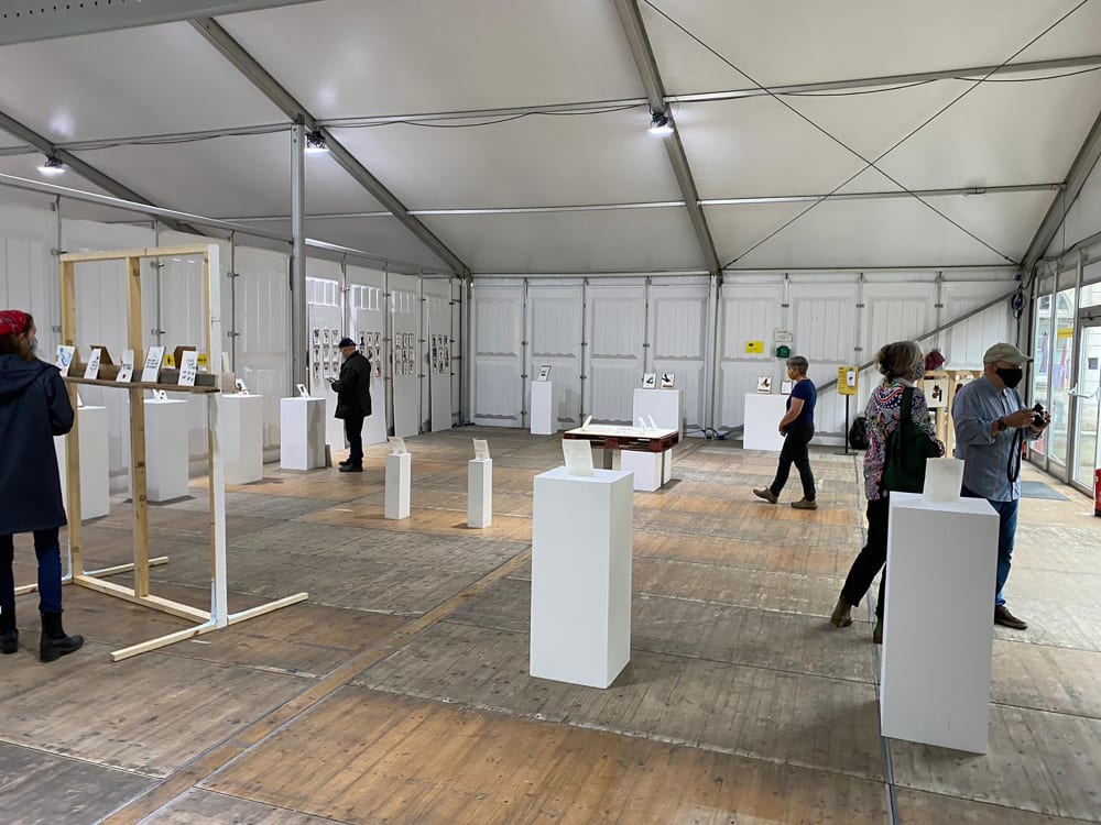 Image is of inside the Chelsea Tent, there are a number of white podiums each with a portrait positioned upright. A number of visitors are walking around the exhibition looking at the podiums.