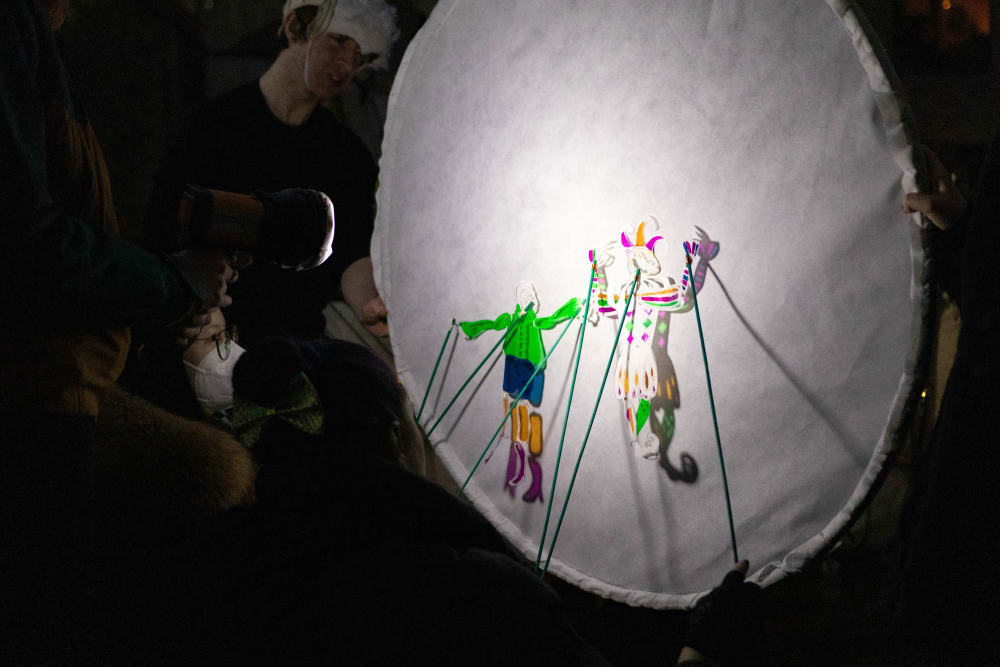 two colourful shadow puppets are held up with sticks to a white circular screen in an otherwise dark space.