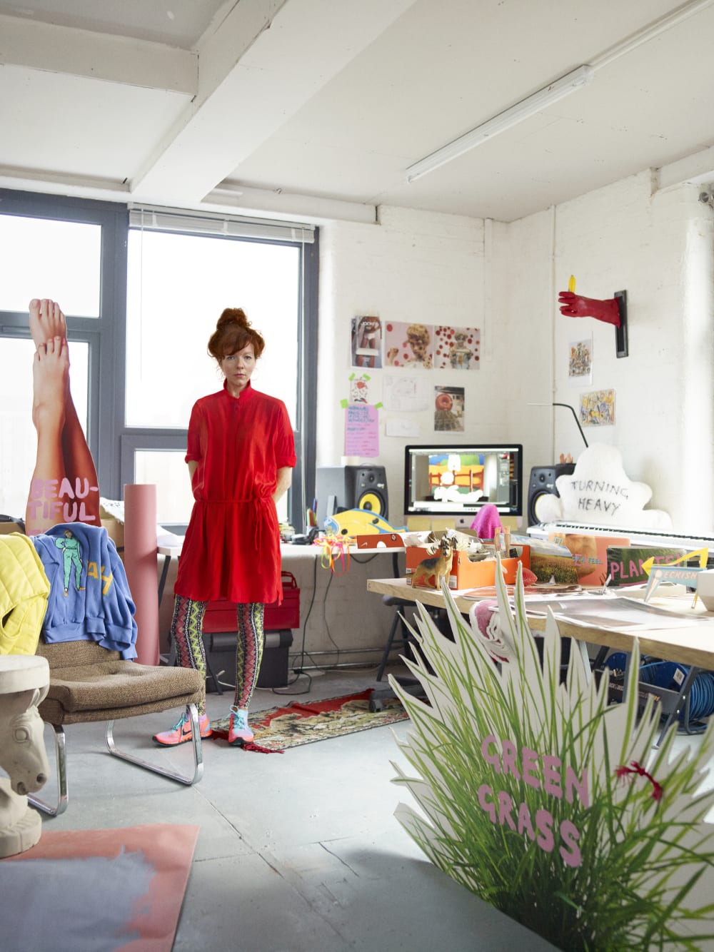 Heather Phillipson standing the studio surrounded by art and wearing a red dress.