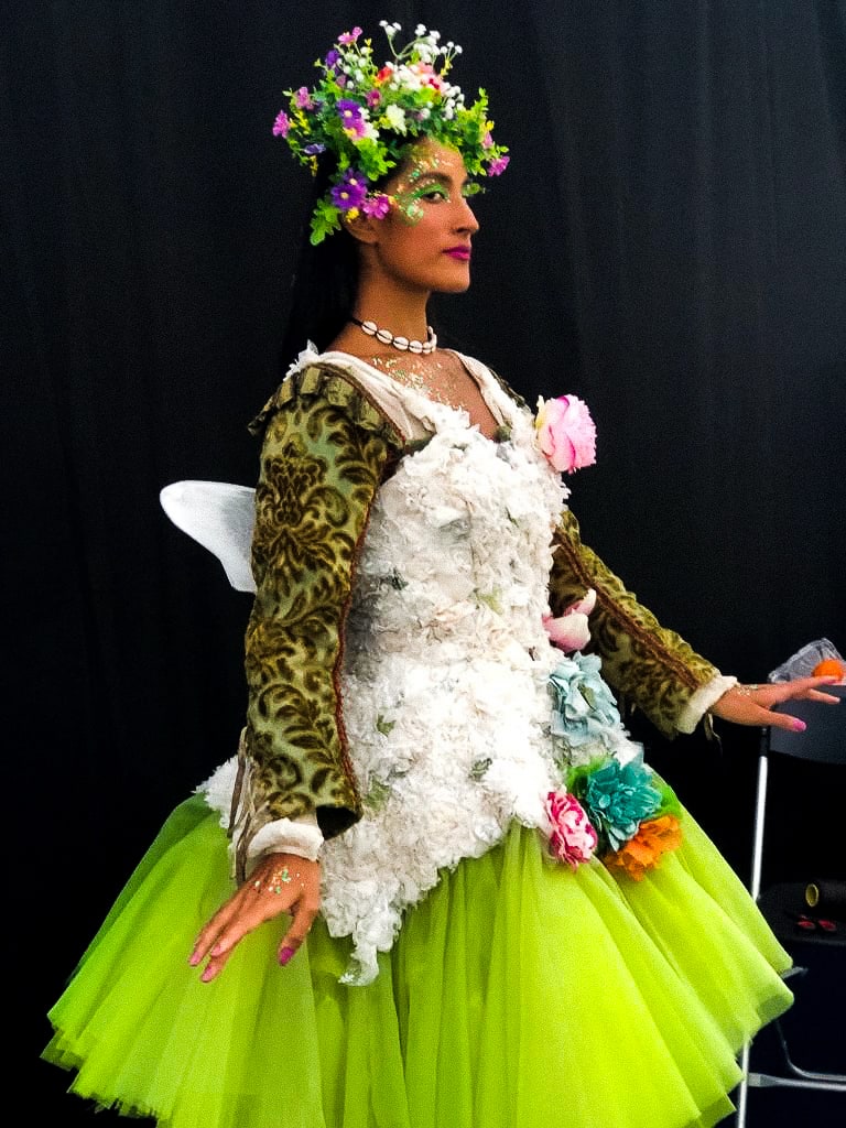 A performer poses in profile in a bright green fairy costume. On her head she wears a garland of flowers 