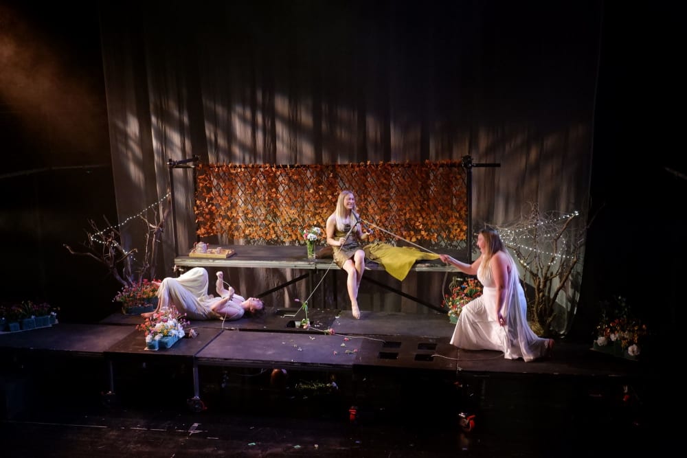 3 women on a stage sitting and reclining wearing togas in what appears to be a garden scene.