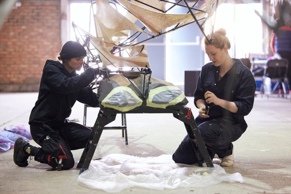 2 puppet technicians are crouched down and working on either side of a component of a puppet 