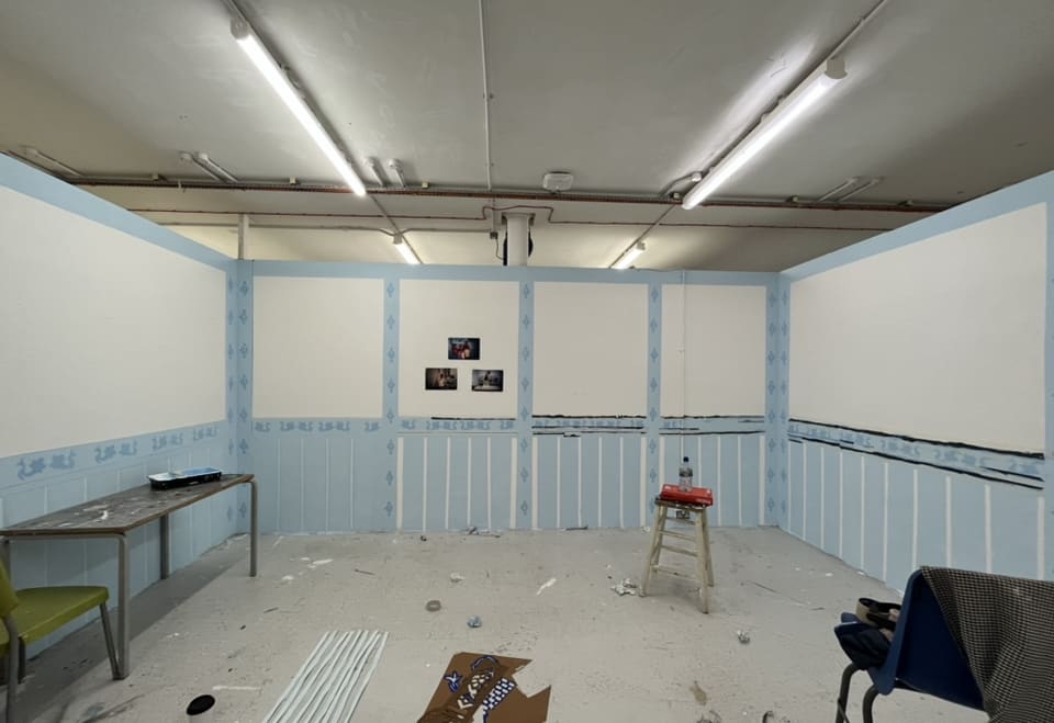 A work in progress installation shot, there are 3 walls in position, painted decoratively in blue and white. Within the installation there is a table with a roller paint tray, a stall with a book and a bottle of water on top and blue strips of white paper are positioned on the floor.