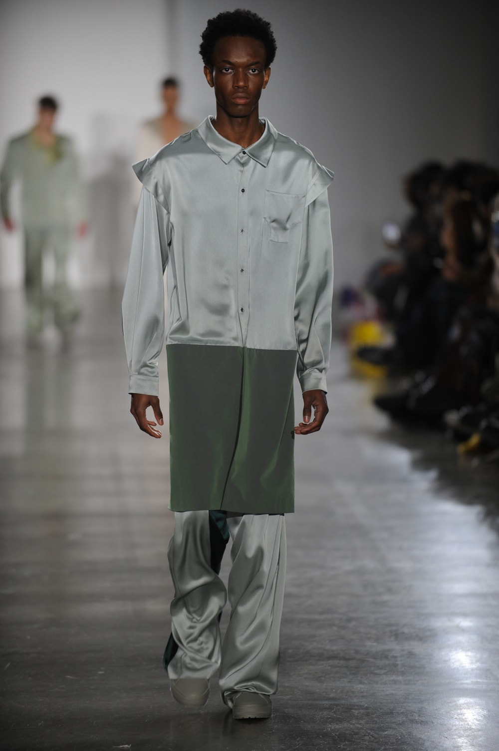 Man on runway wearing silver / green hue buttoned collared shirt with shoulder detailing and left breast pocket, long green hem attached to the bottom of shirt paired with silk straight bottoms with forest green inner and outer leg panel and sage green shoes 