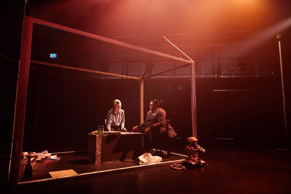 Image shows a dark stage with 2 people sitting at a table in a frame of a house. They appear to be engaged in discussion.