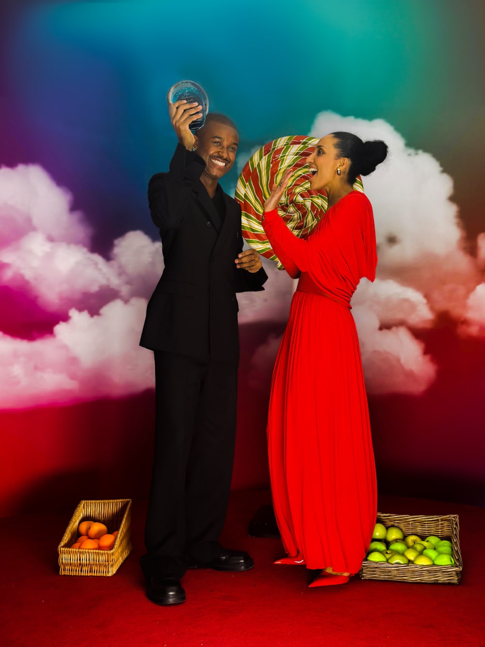 Maximilian and Chioma standing in front of a high-contrast background. Maximilian is holding his award and smiling to the camera; Chioma is looking at him. She's wearing a bright red floor length dress. There's a giant lollipop