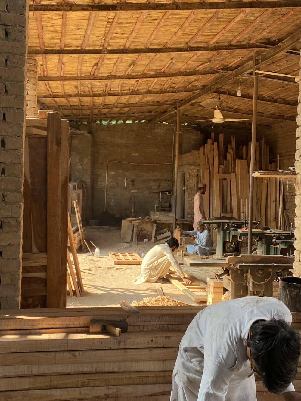 The interior of a workshop in Shikarpur, with pieces of wood, machinery and 4 craftspeople. The sun beams into the bright timber workshop.  
