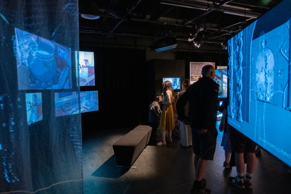 Image shows a dark studio room with a blue screen on the right and blue lit fabric on the left. People can be seen interacting with the screens.