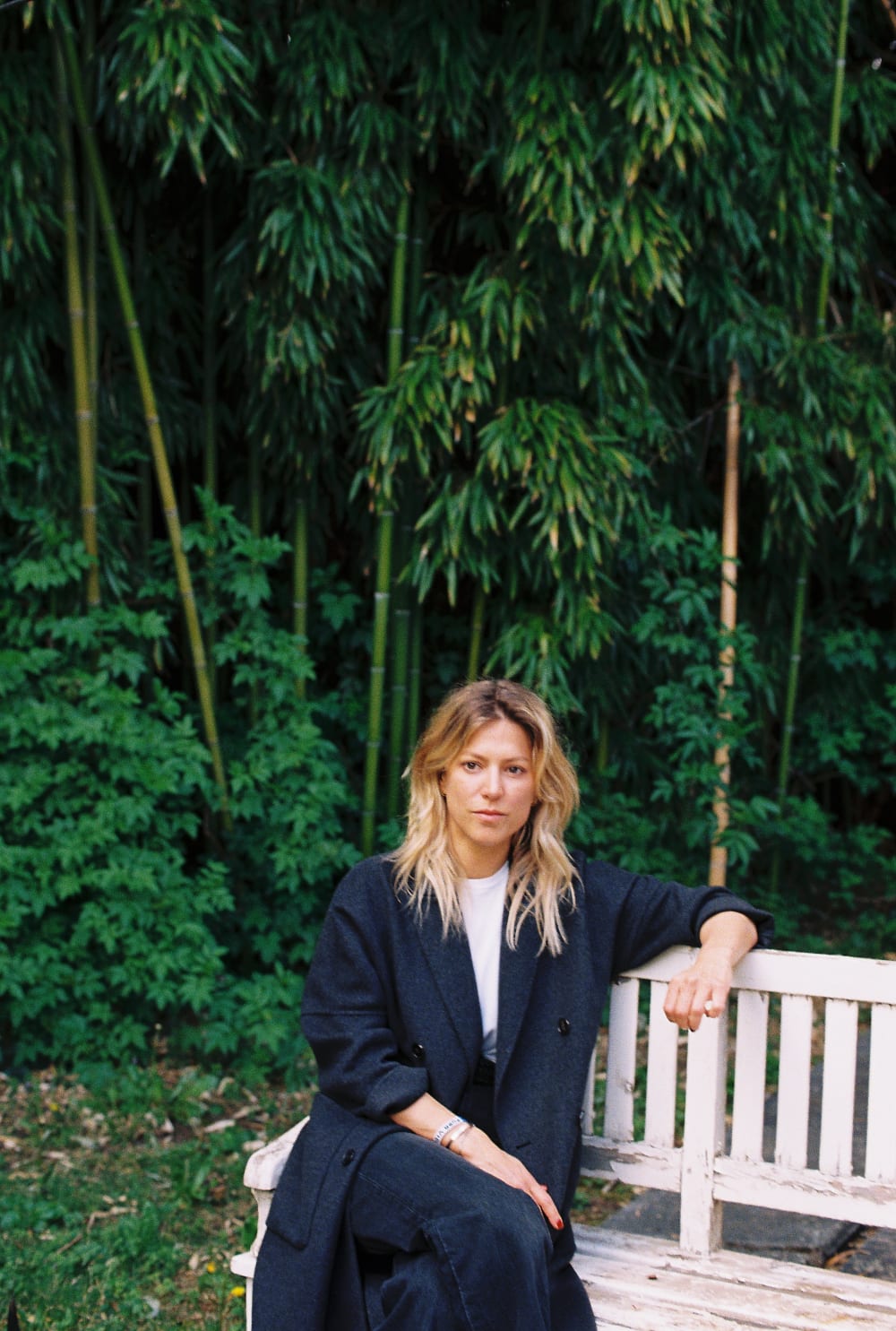 Anna sat on a bench outside. Her left elbow is resting on the back of the bench and she's looking directly into the camera. 
