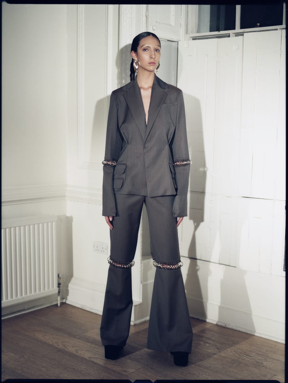 Model wearing a grey, tailored suit with metal detailing around the knees and elbows. The model is standing in a white room, with shuttered windows behind her. 