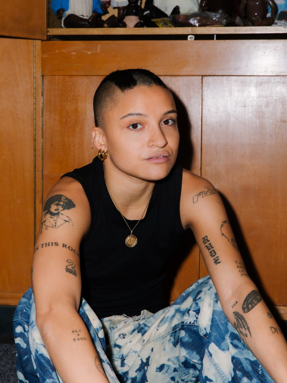 Rene sat on the floor, looking straight into the camera. They're in a relaxed position, with their arms over their knees. They're wearing a black vest and blue acid wash jeans, with a number of tattoos on their arms. 