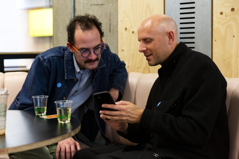 2 men sitting at a table looking at a phone. There's a table in front of the with 2 glasses of white wine on them