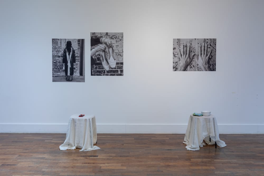 3 black and white photos hanging on a white wall, with 2 small side tables covered in white cloth placed in front. On the table there're pens and henna.