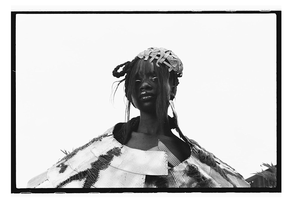 A mono framed shot of a model wearing a criss cross headpiece and matching sculptural outfit featuringa  large, circular layers of brown coffee cup pieces, forming a dramatic collar and shoulder structure that fans outward, with bold black stitching creating striking running patterns. 