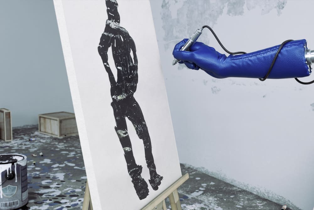 In an art studio, a robotic arm with blue glove is spray-painting a human-shaped figure in black on to a canvas. White paint splotches are visible on the floor and a white paint bucket is just visible in the left hand corner.