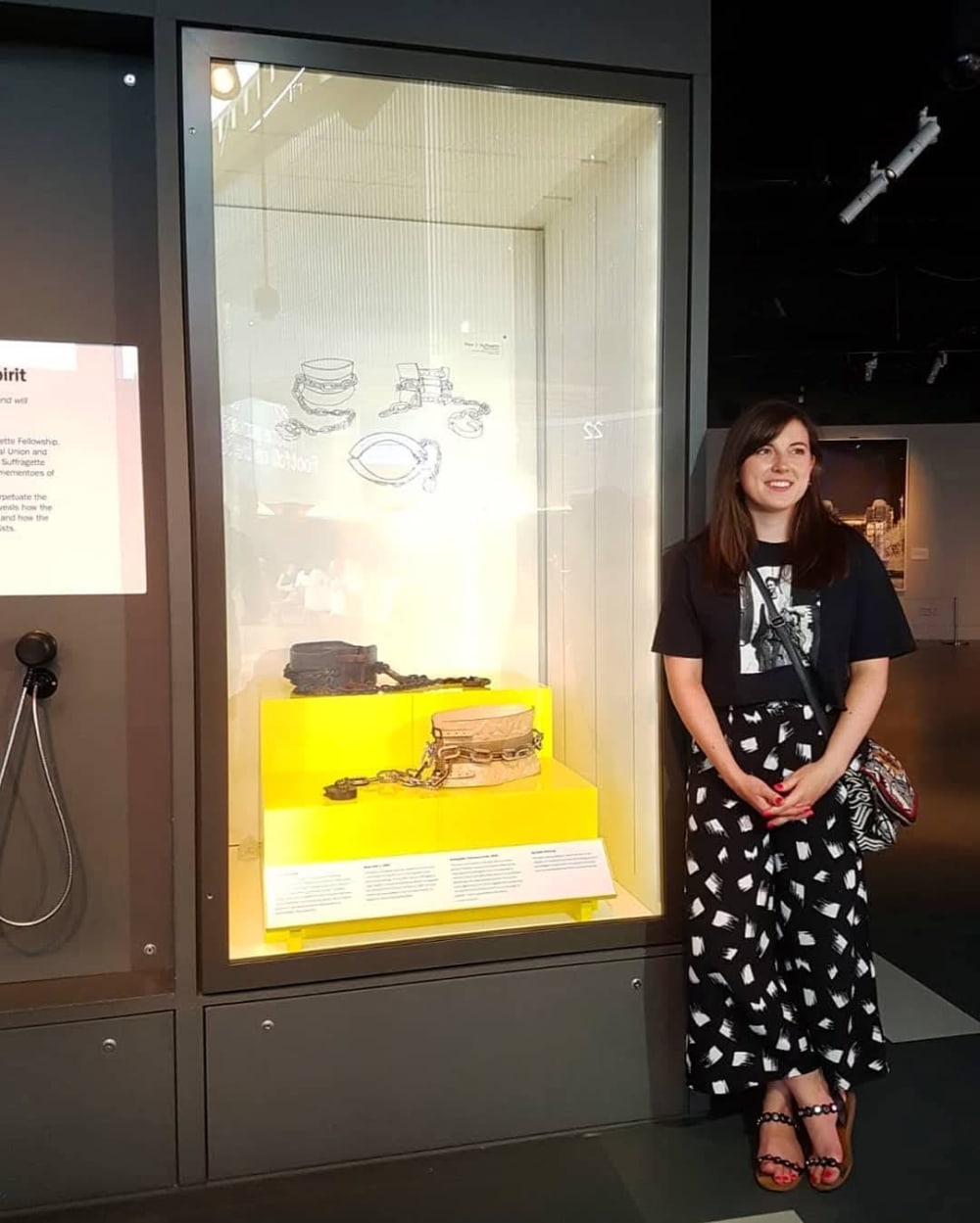 A woman stands smiling next to a museum exhibit displaying historical restraints on a yellow background. The scene conveys a casual, educational atmosphere.
