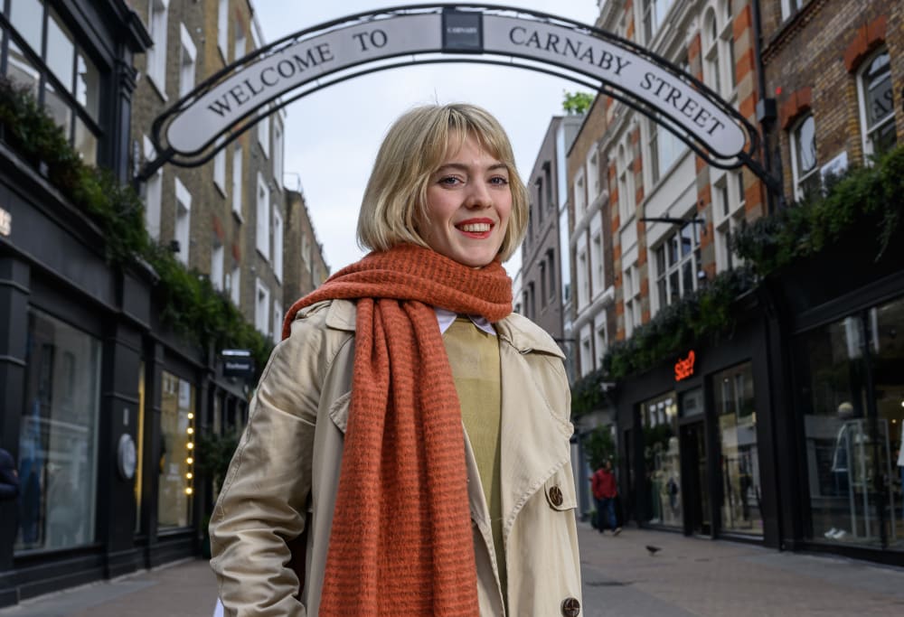 Smiling woman in a beige coat and orange scarf stands on Carnaby Street, under the 