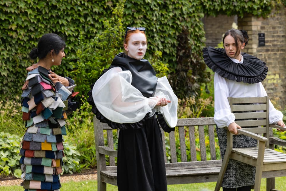  Three individuals wearing creative, sculptural garments stand in a garden area with ivy-covered walls and wooden benches.