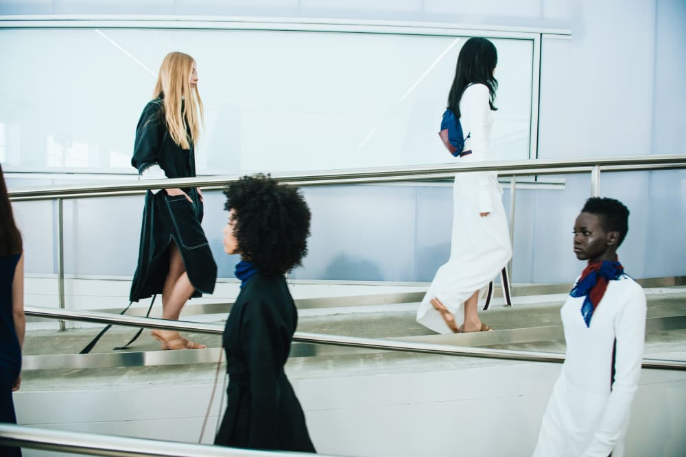 Models pass each other on a sloped walkway. They are wearing black and white garments, some wear navy and red accessories.