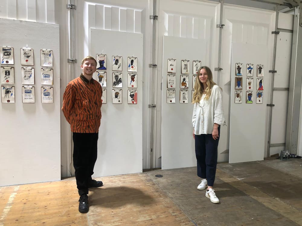 Two people a man in an orange and black striped top, black trousers, black shoes and short hair. A woman with long blonde hair, white shirt black trousers and white trainers. They stand in front of a display of portraits by artist Stephen Anthony David and are looking directly at the camera