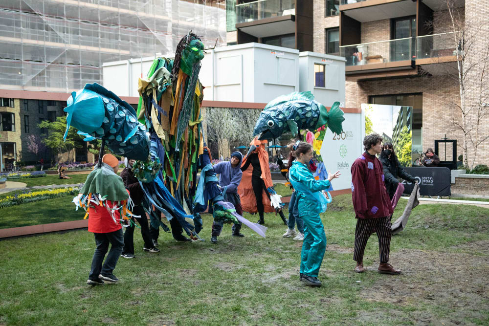 A group of people in colourful outfits operate large puppets of two fish and a green man in an outdoor space.