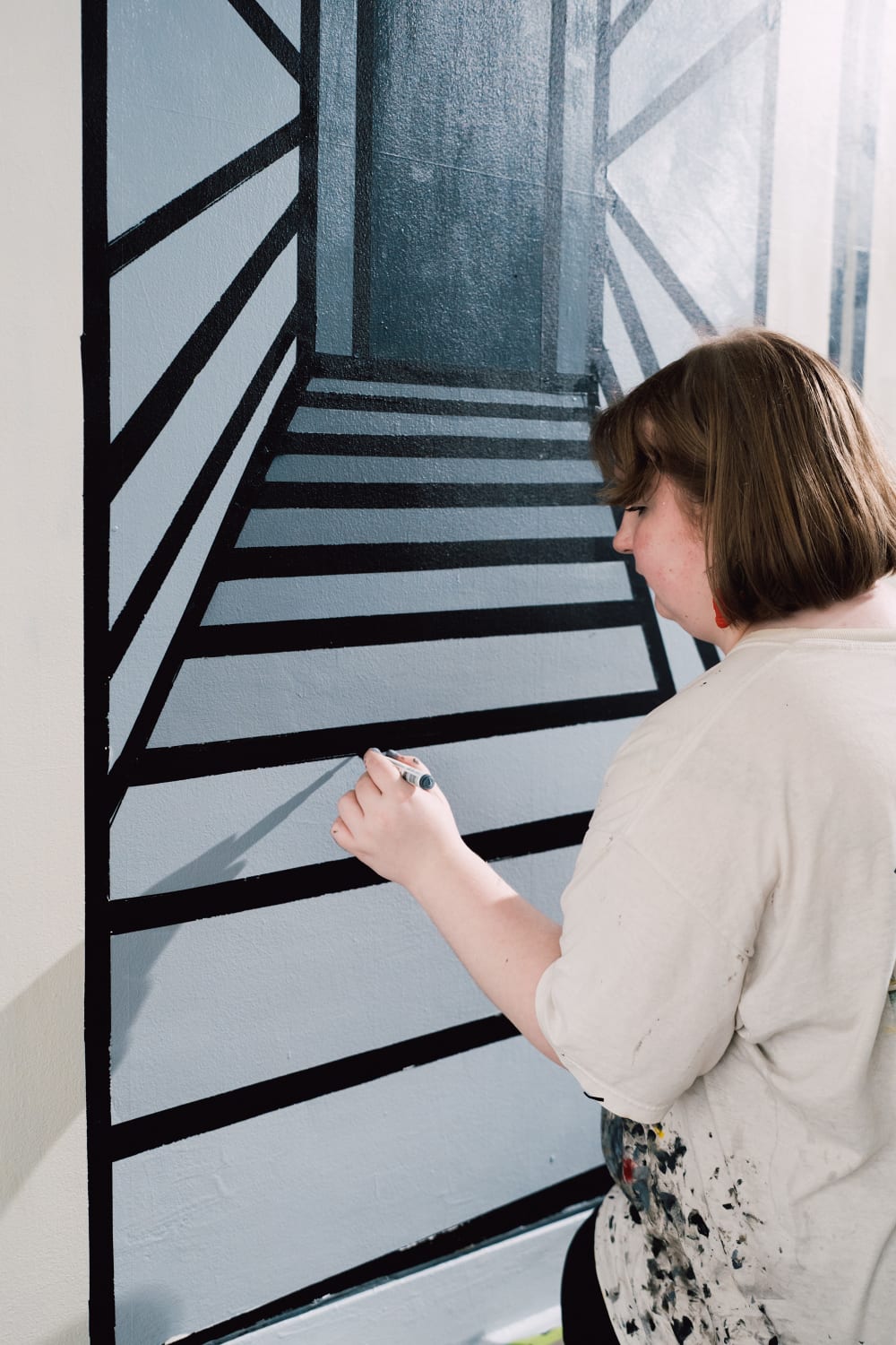 Image shows the side profile of a person in a white top and short brown hair holding a pen and reaching to their left hand side to draw a line on a wall. The wall itself contains a half-completed artwork using different shades of grey paint