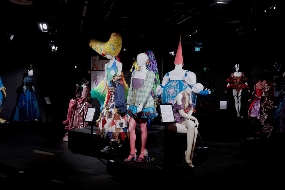 Image shows mannequins standing in a dark room. The mannequin in the centre is wearing a blue checked costume with pink tights. On either side, mannequins can be seen wearing elaborate headwear.