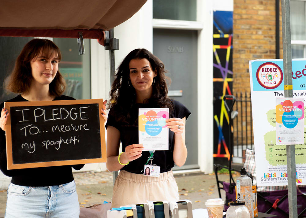 Two girls standing at a stall - one holds a sign that says 'I pledge to measure my spaghetti'