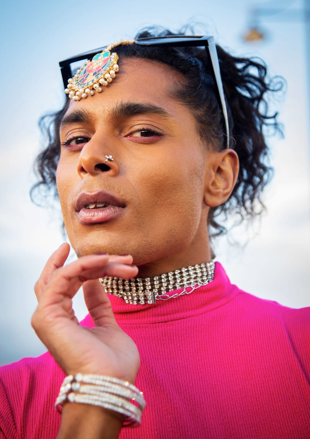Close-up photo taken from below. The person has their hair tied back and is wearing a neon pink, high-necked top; a multi-row diamanté necklace and sunglasses on their head. Their hand is resting under their chin and they're looking directly into the camera
