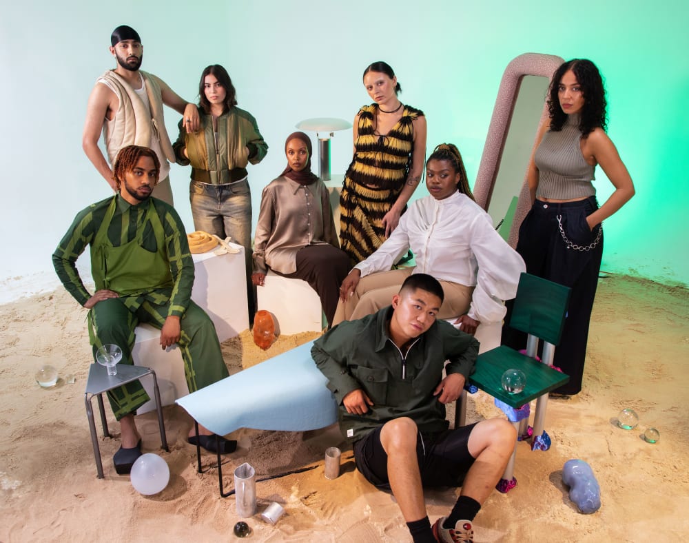 Eight people posing in a studio. The floor is covered in sand and there are different objects dotted around. 
