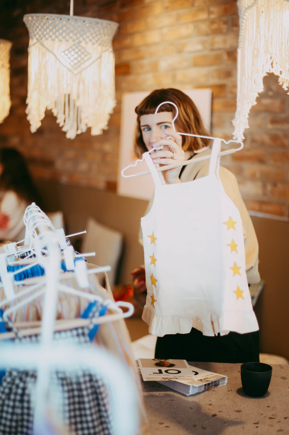 Bliss holding up a white top for a child with yellow embroidery in front of a rail of other clothing