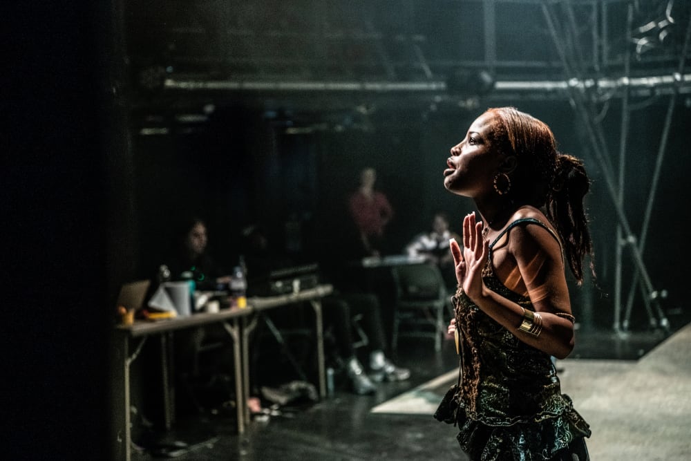 Image shows a young person performing on stage from the waist-up. They are looking up and are wearing earthy colours of brown, green and black.
