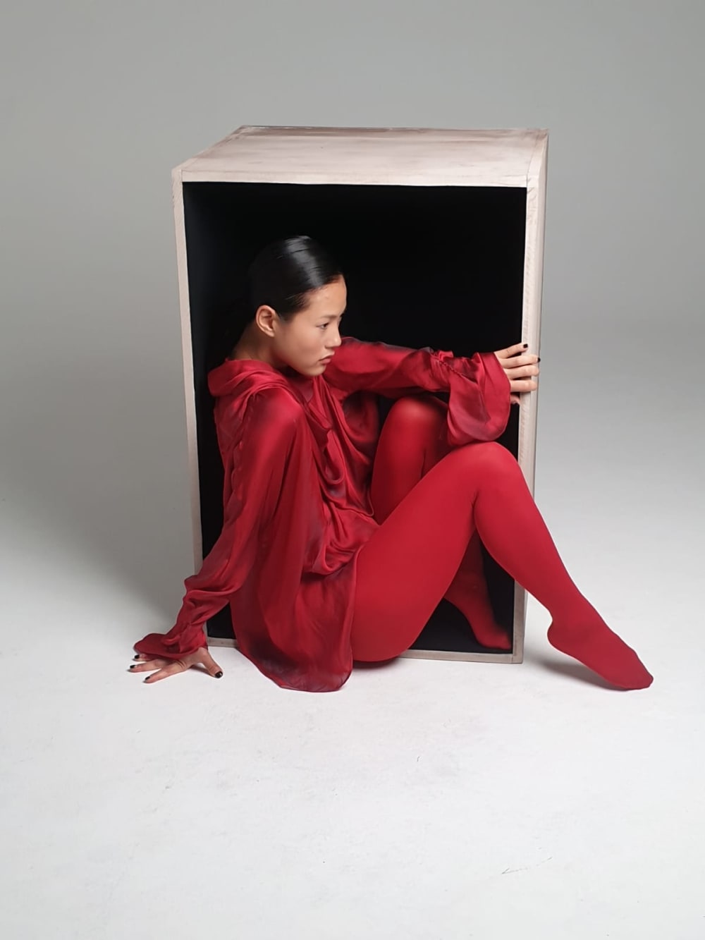 A model wearing a silk red shirt dress and red tights, climbing out of a whitewashed wooden box, lying open on its side, against a white backdrop. 