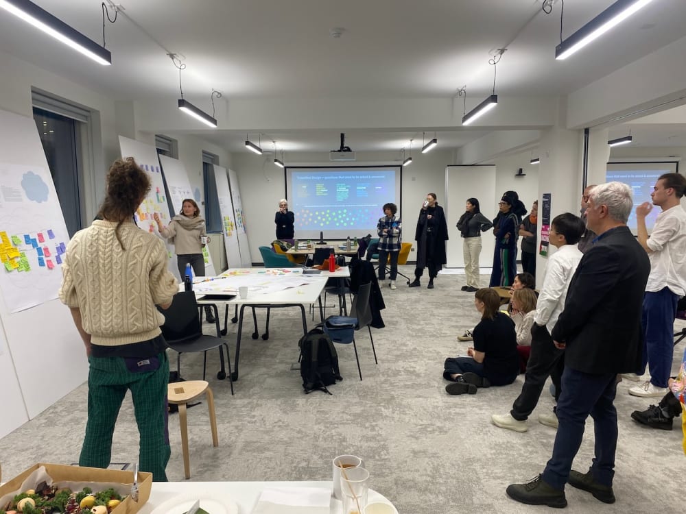 A group of people engaged in a collaborative workshop or presentation in a modern meeting room. The room features large posters with colorful sticky notes and a projector displaying a slide titled 
