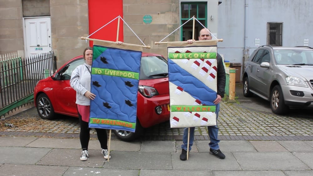 2 people standing outside, holding quilted signs. The sign on the left says 'To Liverpool, to Birkenhead', and the sign on the right says 'Welcome Welcome'.  