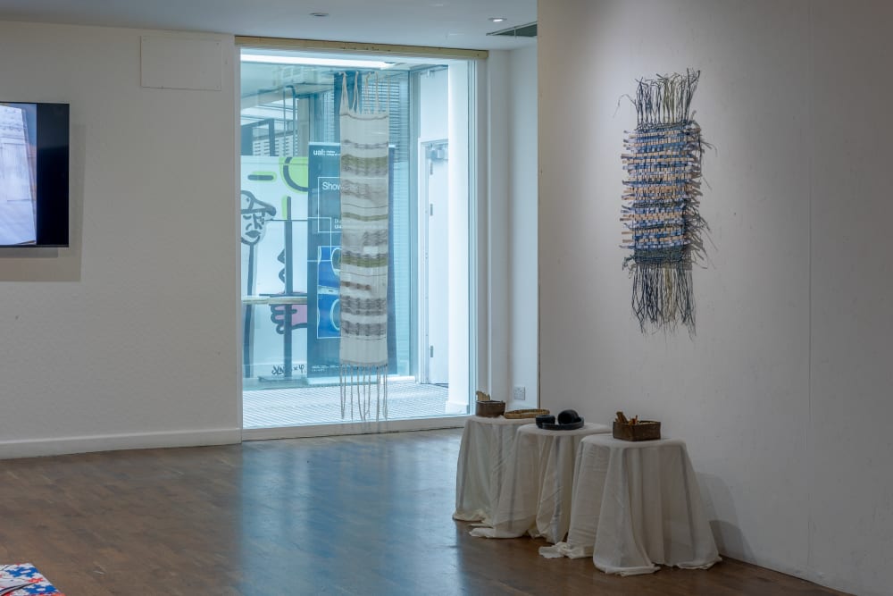 2 woven pieces hanging on the wall. There's 3 side tables in a row to the right, all covered in white material with small bowls on top.  
