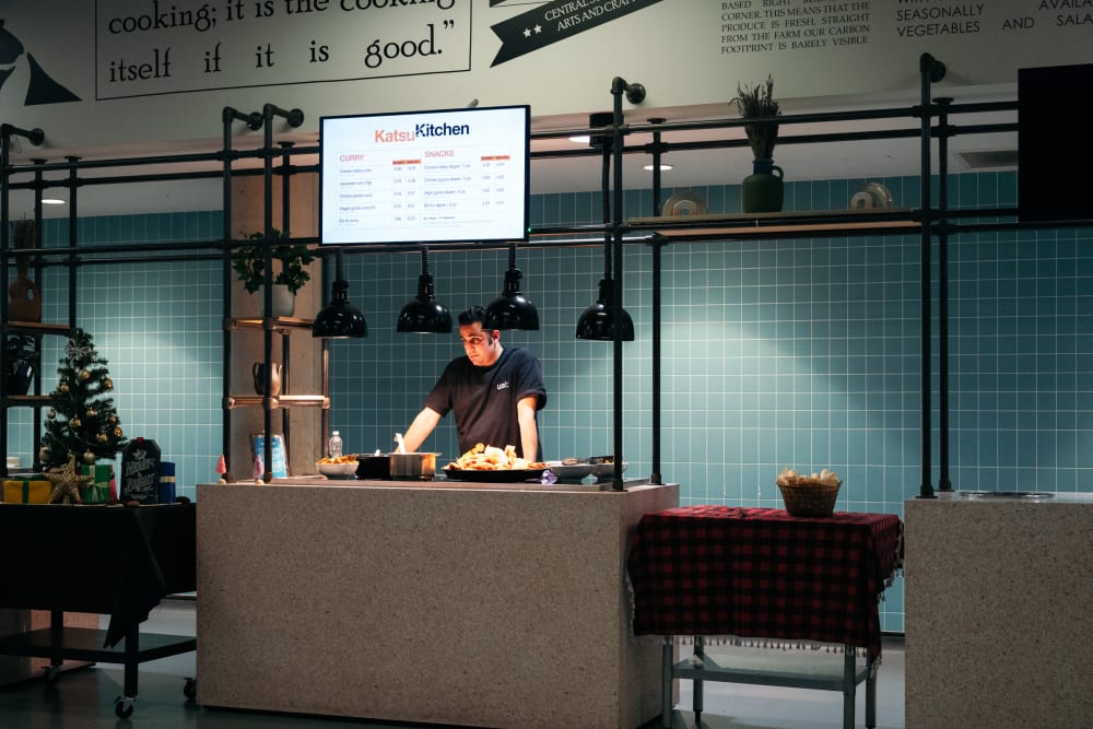 An industrial style canteen counter with a screen above reading 'Katsu Kitchen' and showing a menu. A staff member in a black t-shirt is waiting to serve students and bowls of food are visible on the countertop under heatlamps. The wall behind has square teal tiles and other serving stations are visible to the right and left.