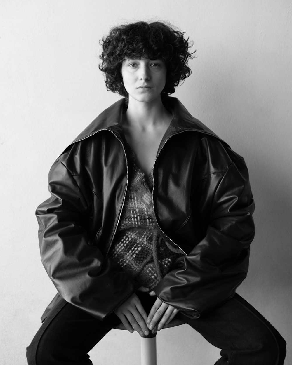 Black and white photo of a model wearing a leather jacket and sitting on a chair, with a white background