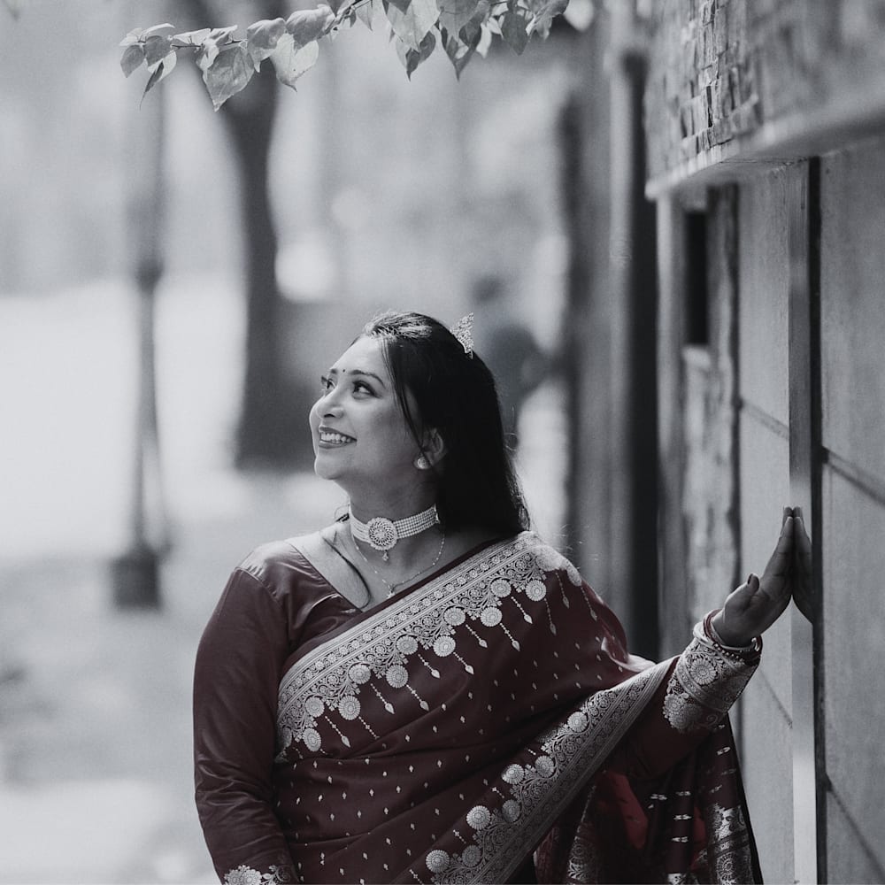 A portrait-style photograph of a person standing beside a stone wall in an outdoor walkway. The person wears a patterned sari with ornate borders and jewelry, with one hand resting on the wall and the head turned upward toward overhanging leaves. The image uses selective color, highlighting the deep red tones of the sari while the background and most other elements appear in black and white. The background is softly blurred, showing trees and architectural features along the path.
