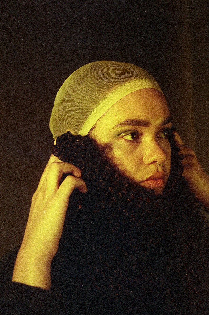 profile of a female with an artificial hair piece she is holding up with both hands as a beard